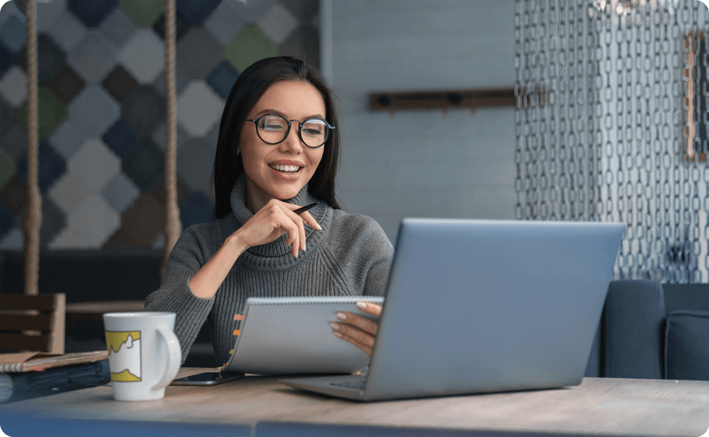 Professional woman wearing glasses, working at a desk with a laptop and notepad, smiling as she reviews information.